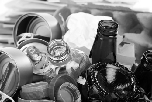 The image depicts a collection of various used beverage containers and waste items, including several aluminum cans with pull tabs and open tops, some of which are crushed or deformed. There are also clear glass jars with threaded necks, one upright and another lying on its side, alongside black glass bottles, possibly empty. Nearby, a black leather handbag with stitched detailing and metal hardware is partially visible, resting among the waste materials. The objects are arranged on a surface that appears to be a kitchen or garden waste collection area in Redbridge, with some paper or tissue material in the background. The lighting is natural, providing clear visibility of the different textures and colors, mainly metallic, glass, and leather. This scene illustrates typical domestic rubbish that Garden Clearance Redbridge would handle for efficient removal, in an environment that suggests preparation for disposal or recycling, consistent with their garden and household waste clearance services in the local area.