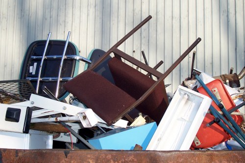 A large pile of discarded household and garden items arranged outdoors on a weathered surface, including a brown fabric-upholstered chair with wooden legs tilted sideways, a blue plastic storage bin partially visible, a white door or panel leaning against the pile, and various metal and plastic objects such as red and blue crates, a black wire basket, and parts of a small white appliance. The background features a corrugated metal wall with a weathered finish, indicative of a storage or waste disposal area. The collection of rubbish appears to be in the process of being removed or cleared, consistent with garden clearance or rubbish removal services in Redbridge or nearby areas. The scene is well-lit, with natural daylight illuminating the different textures and colours of the debris, emphasizing the mix of materials including plastic, metal, wood, and fabric, typical of household dump waste that Garden Clearance Redbridge might manage.
