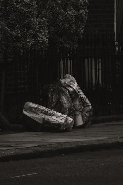 A pile of mixed waste materials including black plastic trash bags, a yellow plastic container, and an upright, worn car seat on the gravel surface of an outdoor area, close to a stone wall. In the ba