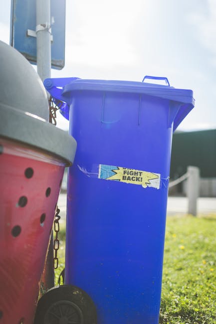 A tall blue plastic rubbish bin with a closed lid is positioned outdoors on a grassy area, with a small yellow sticker on its front that reads 'FIGHT BACK!' in black text with a lightning bolt graphic